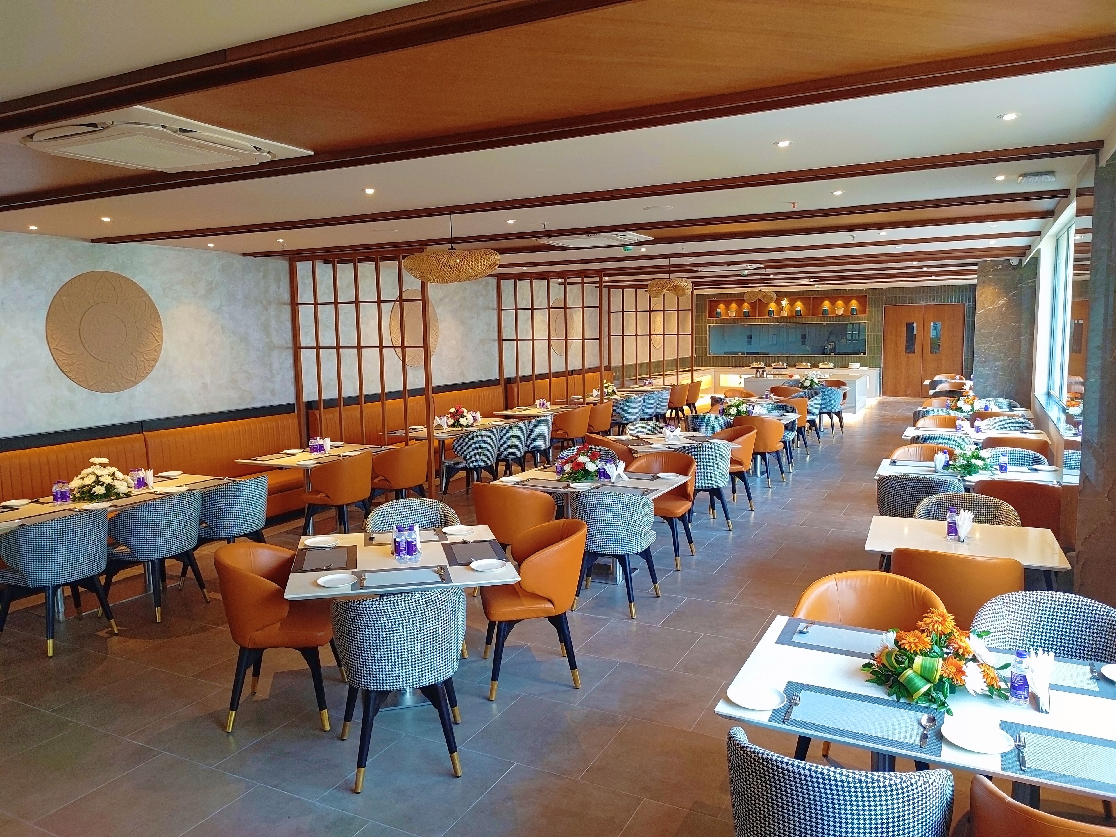 A spacious restaurant with multiple dining tables and chairs, a wooden ceiling, and a view of the kitchen area.