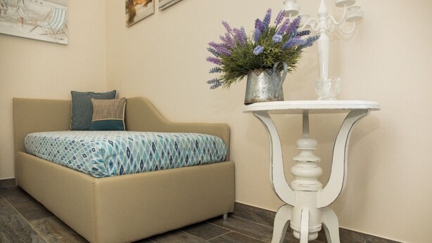 Ein Zimmer mit einer beigen Couch, einer gemusterten Decke, einem kleinen weißen Tisch mit Blumenvase und einer Wandlampe.