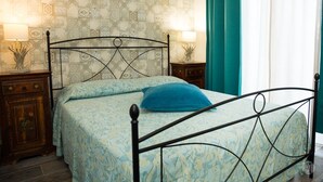 Ein Bett mit Metallgestell, einer gemusterten Tagesdecke und einem blauen Kopfkissen. Zwei Nachttische mit Lampen und dekorativen Vasen stehen bereit. Der Raum ist mit gemustertem Tapeten und lindgrünen Vorhängen gestaltet.