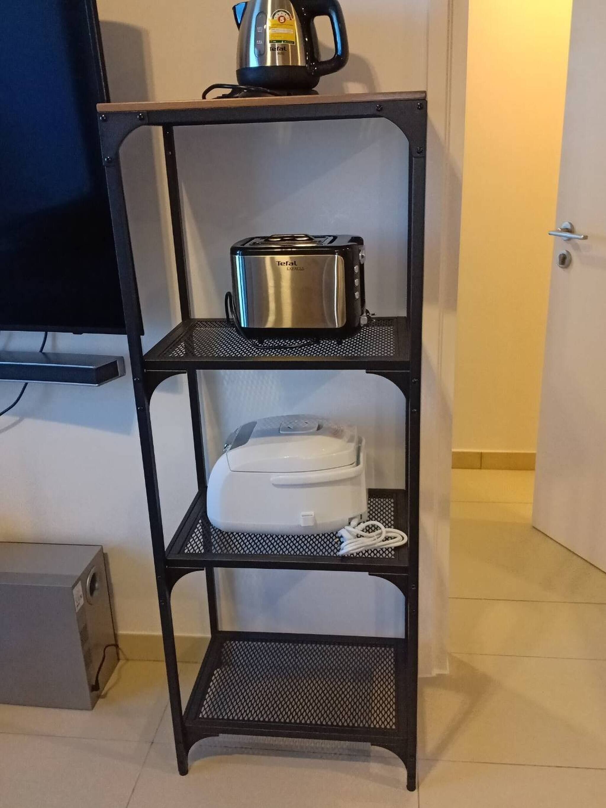 A black metal shelf unit with a toaster and a kettle on the top shelf, a white appliance on the middle shelf, and a white device on the bottom shelf.