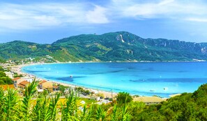 Uma cidade costeira com uma praia, águas azuis cristalinas e colinas verdejantes ao fundo.