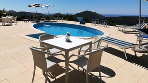 Piscina com espreguiçadeiras e mesas, rodeada por montanhas.