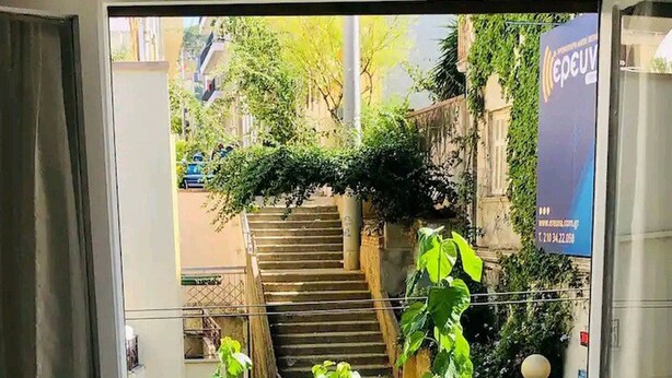 Ein offener Balkon mit einem Holztisch und Stühlen, Blick auf eine Treppe und Grünflächen, und ein Banner mit Schrift.