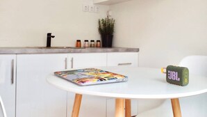Eine moderne Küche mit weißen Schränken, einer weißen Arbeitsplatte und einem runden Tisch mit Stühlen. Darüber befinden sich Regale mit Geschirr und Gläsern, und auf dem Regal steht eine kleine Pflanze.