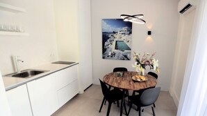 A modern kitchen with white cabinets, a stainless steel sink, and a wooden dining table with black chairs. There is a painting of a coastal scene on the wall and a vase with flowers on the table. - Junior Unicorn Suite (Athens)