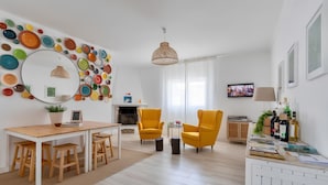 A modern living room with a white fireplace, yellow armchairs, a dining table with wooden stools, a colorful circular wall decoration, and a television. - Galagana Charm House (Golega)
