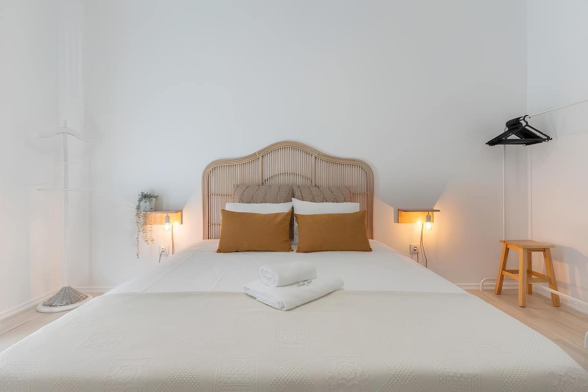 Ein ordentlich gemachtes Bett mit weißer Daunendecke und zwei braunen Kissen, einem Holz-Kopfende, Nachttischlampen, einem kleinen hölzernen Hocker und einem schwarzen Kleiderständer.