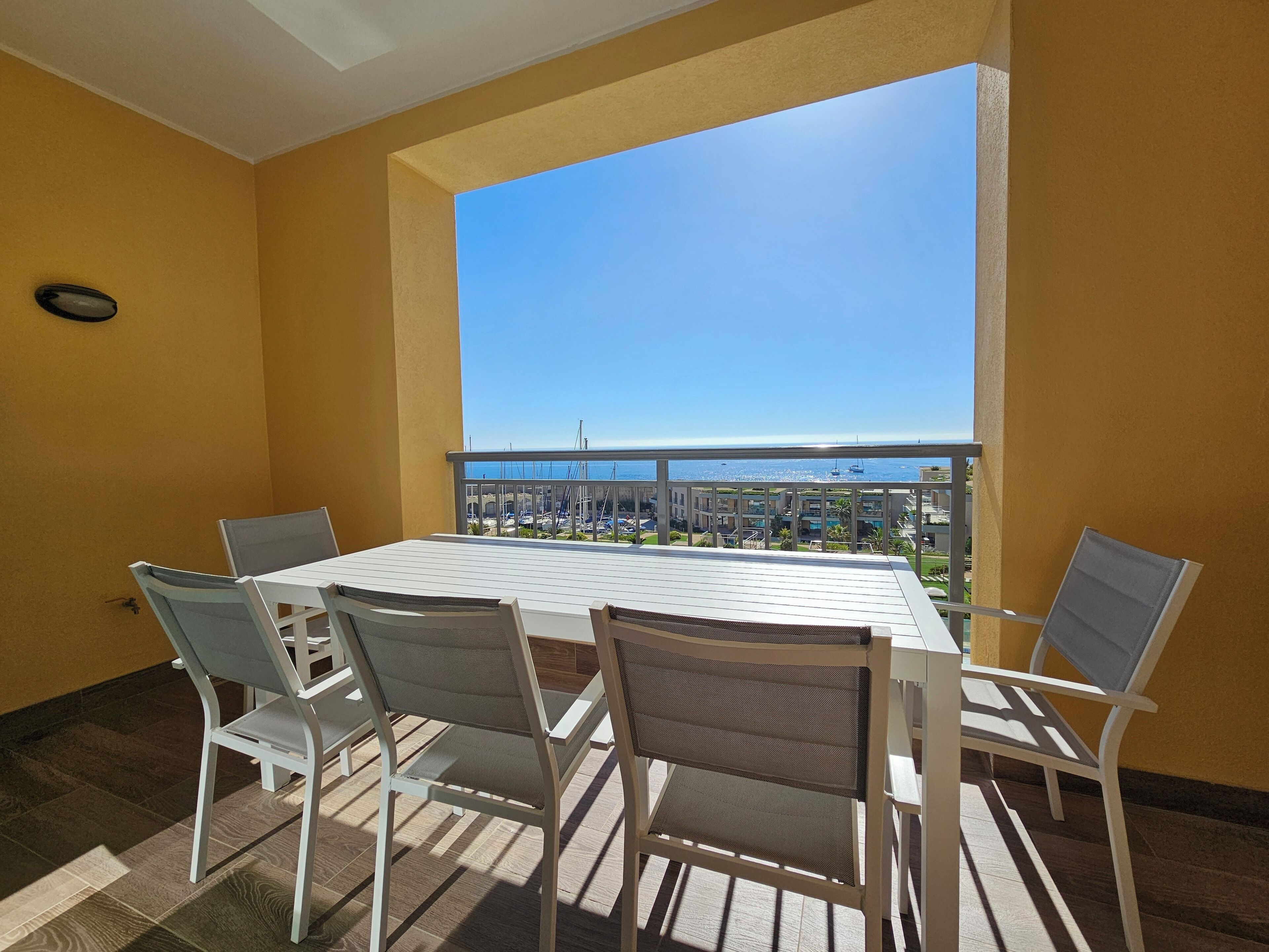 A balcony with a table and chairs, a view of the sea and buildings, and yellow walls.