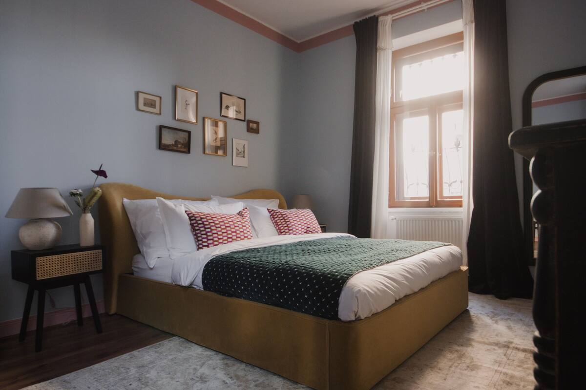 Un dormitorio con cama, una mesita de noche con lámpara, una ventana con cortinas y cuadros en la pared.