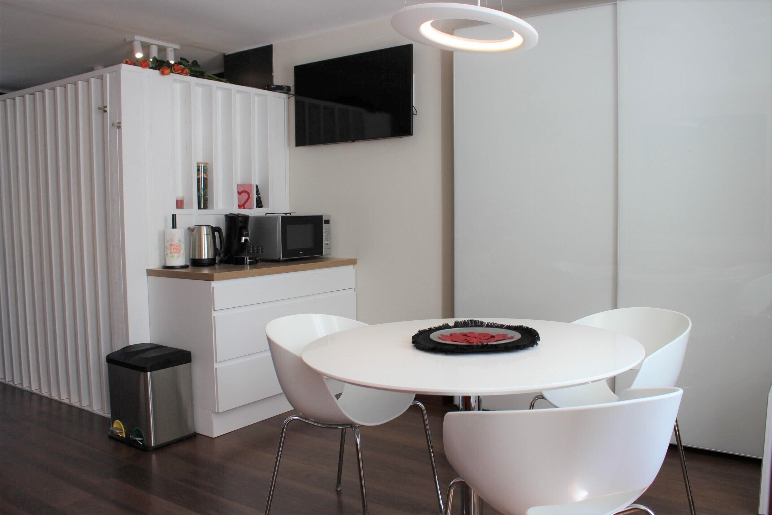 Una cocina moderna con armarios blancos, un microondas y una cafetera. Hay una mesa redonda con sillas blancas y un mantel individual negro.