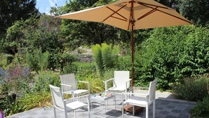 Une terrasse extérieure avec deux chaises blanches et une table, un parasol beige et un jardin avec différentes plantes et fleurs.