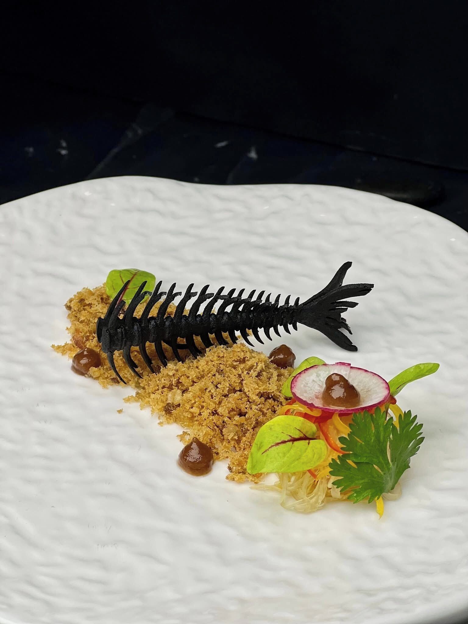 A white plate with a fish-shaped food sculpture, crumbled bread, olives, radish slices, and parsley.