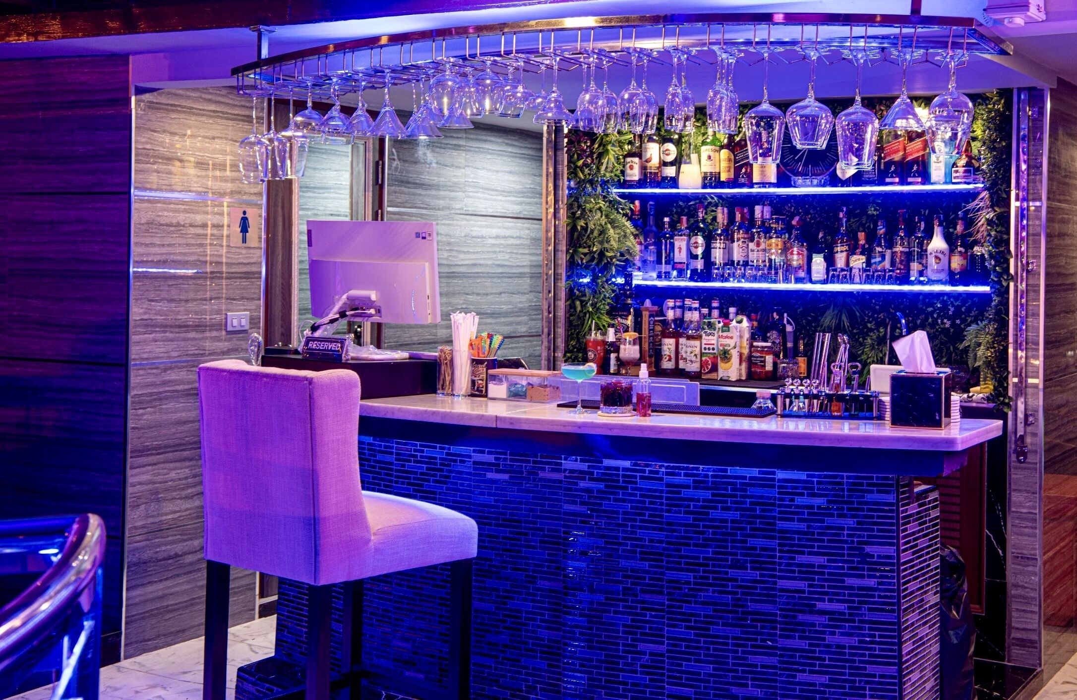 A bar area with a blue-lit counter, a seating area with a chair, and shelves stocked with bottles and glasses.