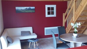 Una habitación con techo de madera, un sofá blanco, una mesita con una silla, una mesa roja y una ventana con persianas.