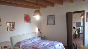 Ein Schlafzimmer mit einem Bett, einem Nachttisch, einer Lampe und einem Sessel. Durch eine offene Tür ist eine Essecke sichtbar.