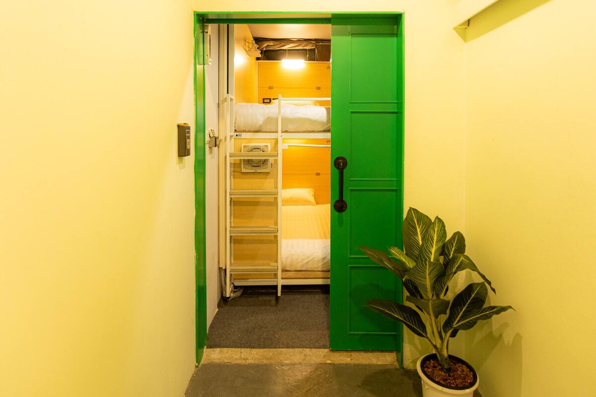 Een smalle gang met een groene deur die leidt naar een kamer met stapelbedden en een plant in pot.