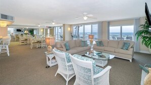 A spacious living area with a large sofa, two white wicker chairs, a glass coffee table, and a dining area with a table and chairs.