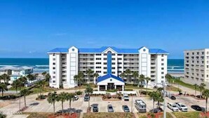 A multi-story hotel with a central entrance, parking lot, and ocean view.