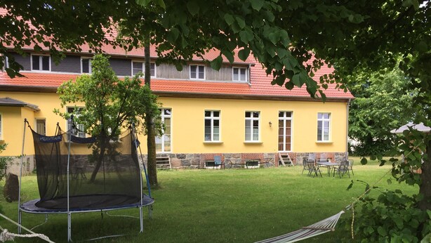 Un bâtiment jaune avec un toit rouge, un trampoline et un hamac dans une cour verdoyante.
