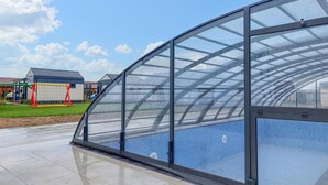 Une structure entièrement vitrée qui offre une vue sur une piscine aux carreaux bleus et sur un espace verdoyant, avec un bâtiment visible au loin.