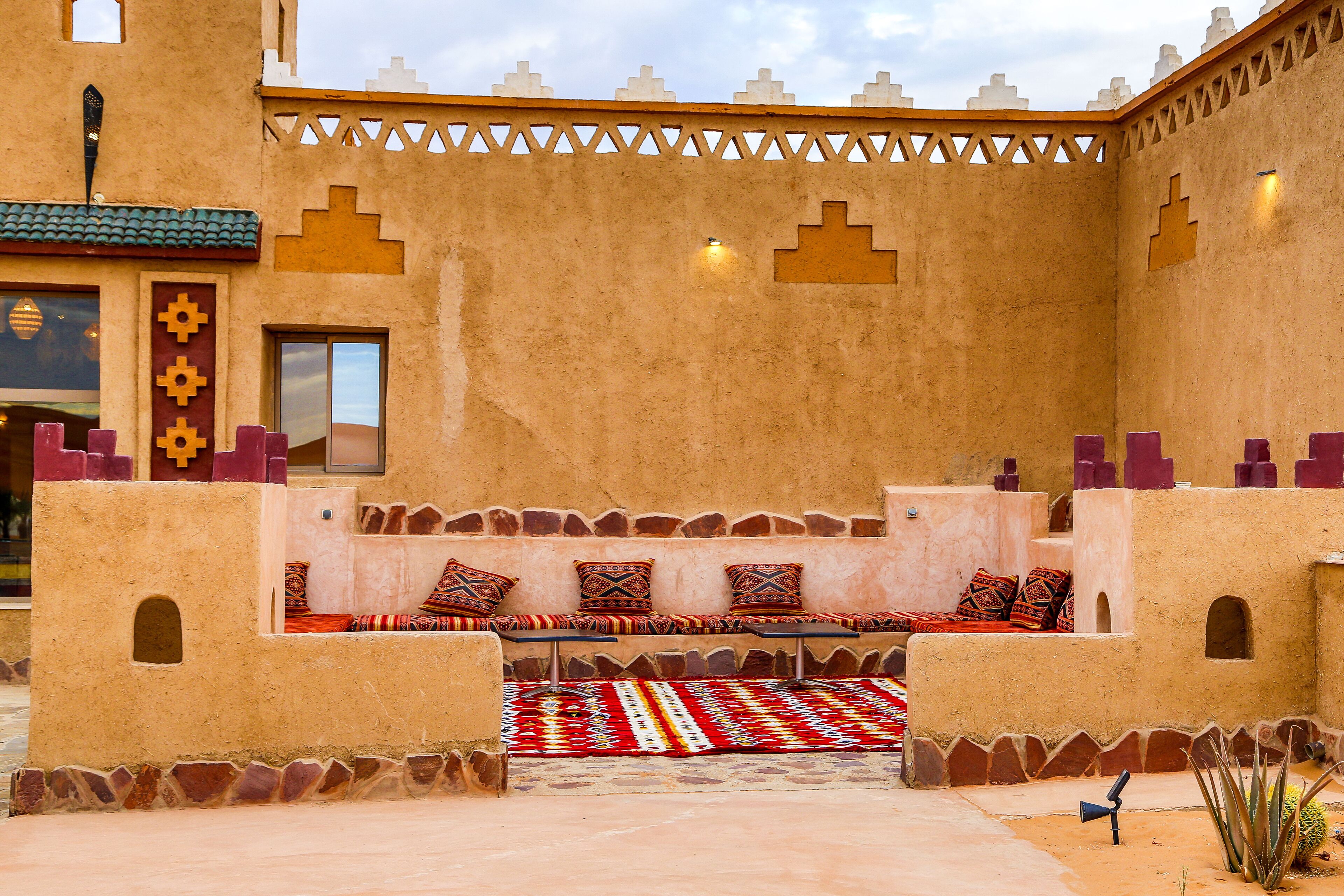 A courtyard with a seating area featuring a patterned rug and cushions, a small table, and a cactus plant.
