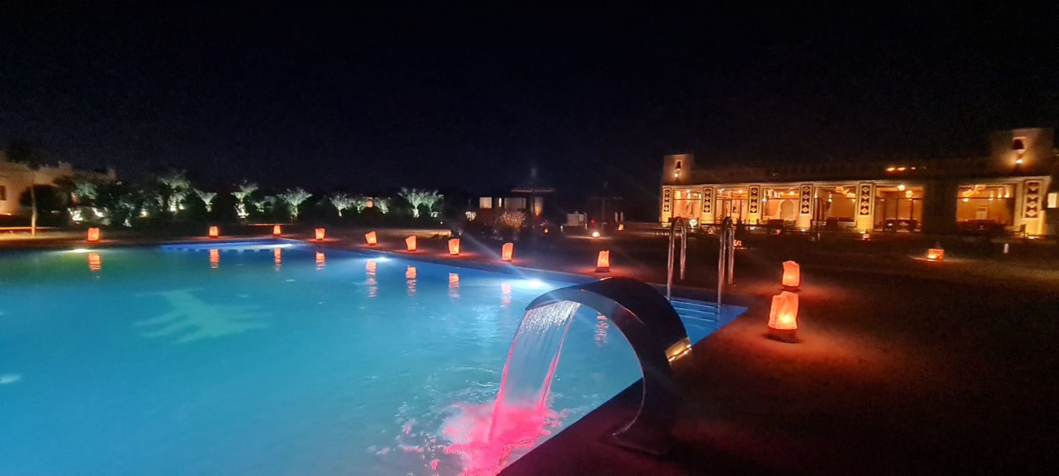 A nighttime swimming pool with water fountains and illuminated by warm lights.