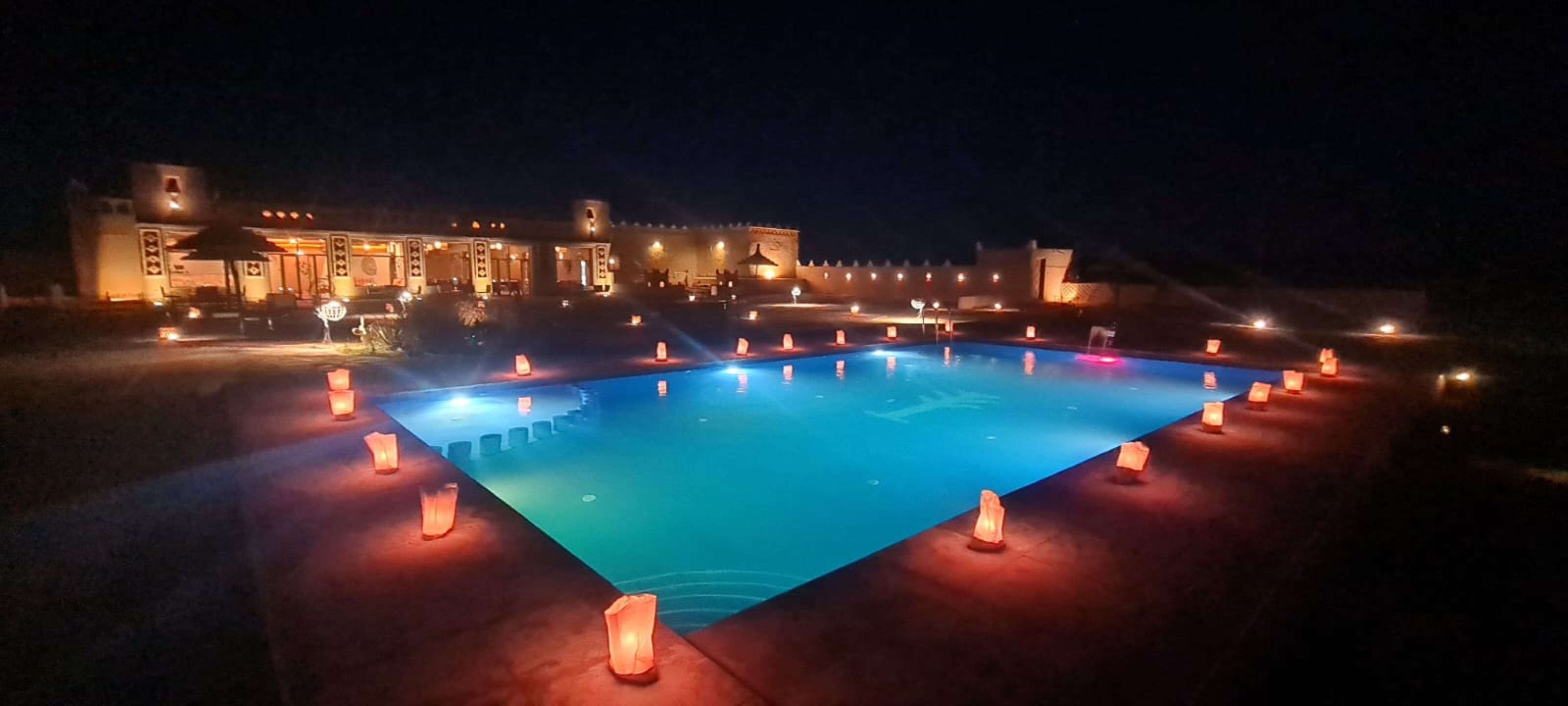 A nighttime swimming pool with illuminated candles around its perimeter, a building with traditional architecture in the background, and a clear sky.