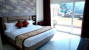 Una cama grande con cabecera, dos almohadas y una colcha. Se ve una silla y una ventana con cortinas.