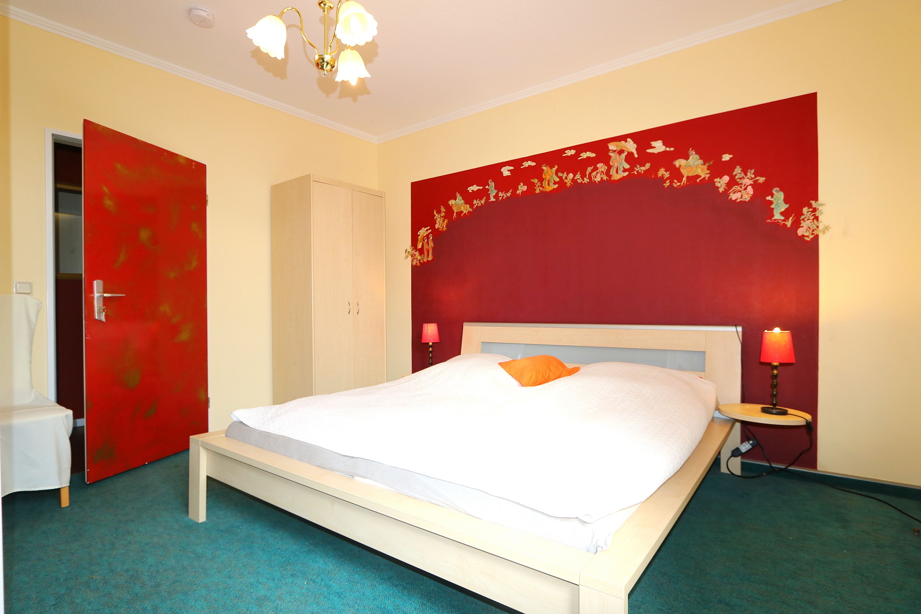 A hotel room with a bed, a red door, a white headboard, and a red wall with a decorative border.
