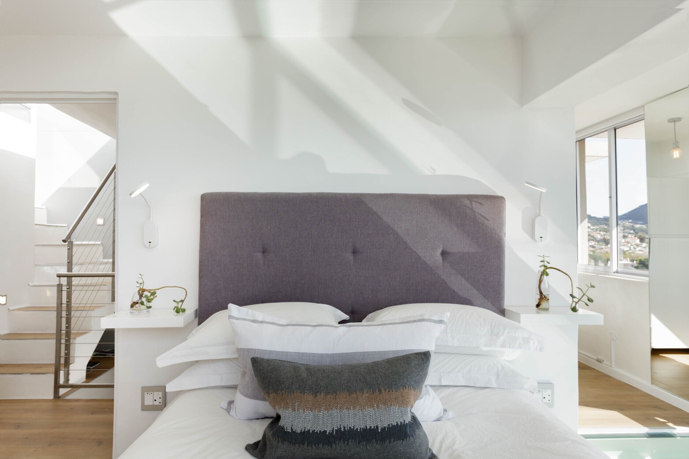 Ein modernes Schlafzimmer mit einem großen Bett, einem grau gepolsterten Kopfteil und Blick auf eine Stadtlandschaft durch große Fenster.