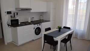Une cuisine compacte avec des meubles blancs, un four à micro-ondes, une cuisinière et un lave-linge. On y trouve une table à manger avec des chaises et une plante en pot.