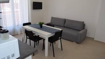 Un ambiente compatto con un tavolo da pranzo e sedie, un divano grigio, una TV a schermo piatto fissata al muro e un angolo cottura con un forno a microonde.