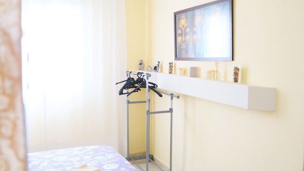Une chambre d’hôtel comprenant un lit, un luminaire suspendu, un portant à vêtements et une étagère avec des objets décoratifs.