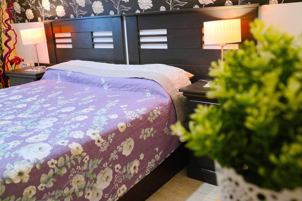 Un llit ben arropat amb un sobrell d'estil floral, un panell de capçal modern amb il·luminació integrada i una taula de nit amb una làmpada.