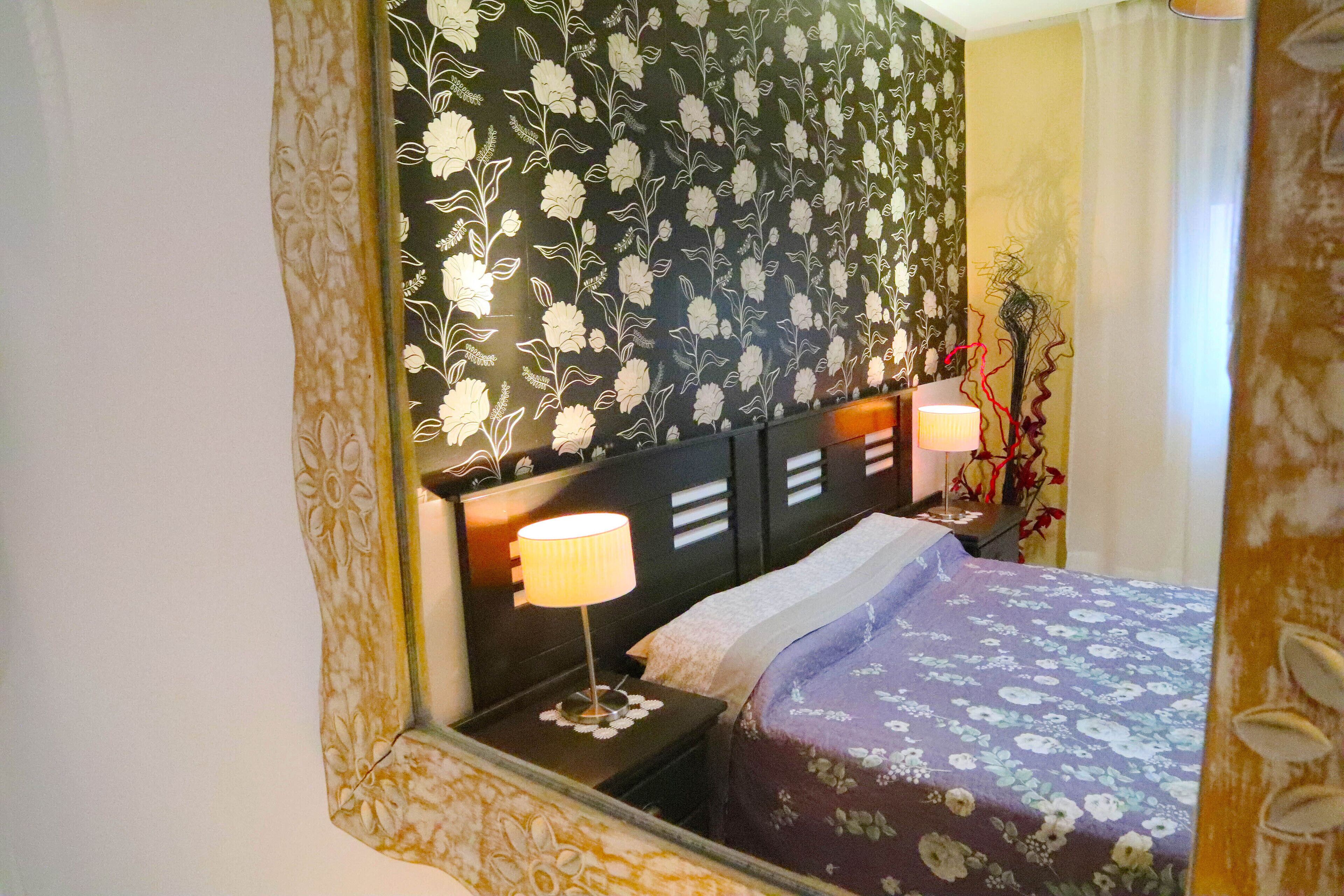 Une chambre d’hôtel avec un lit, des tables de chevet, un miroir et une tête de lit ornée de motifs floraux.
