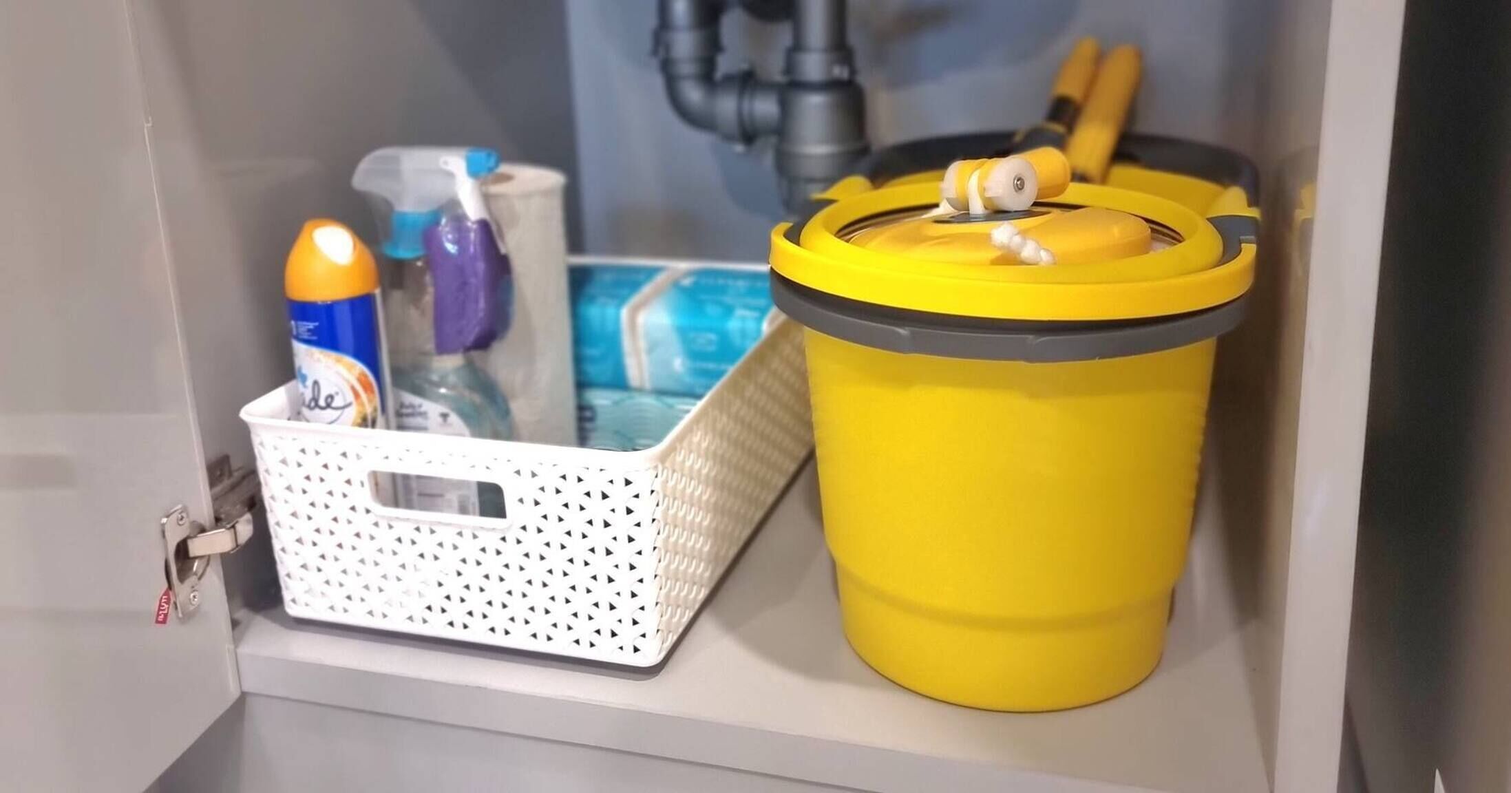 A storage area with a yellow trash bin and a white basket containing cleaning supplies.