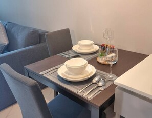 Et spisebord dækket med hvide tallerkner, skåle, bestik og et vinglas, med en sofa og et hvidt skab i baggrunden.