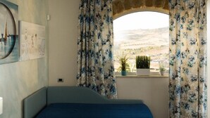 Une chambre à coucher avec un lit, une fenêtre ornée de rideaux à motifs floraux, une vue sur la campagne et un tableau accroché au mur.