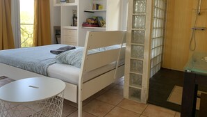 Een slaapkamer met een bed, nachtkastje, inloopkast met glazen deuren, badkamer met douche en raam met gordijnen.