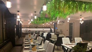 A restaurant with long communal tables, individual booths, hanging plants, pendant lighting, and a visible kitchen area in the background. - Culture by BVH Hospitality Greater Noida (Greater Noida)