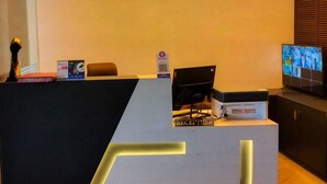 A reception desk with a modern design, a computer, a printer, and a television displaying a video conference. - Culture by BVH Hospitality Greater Noida (Greater Noida)