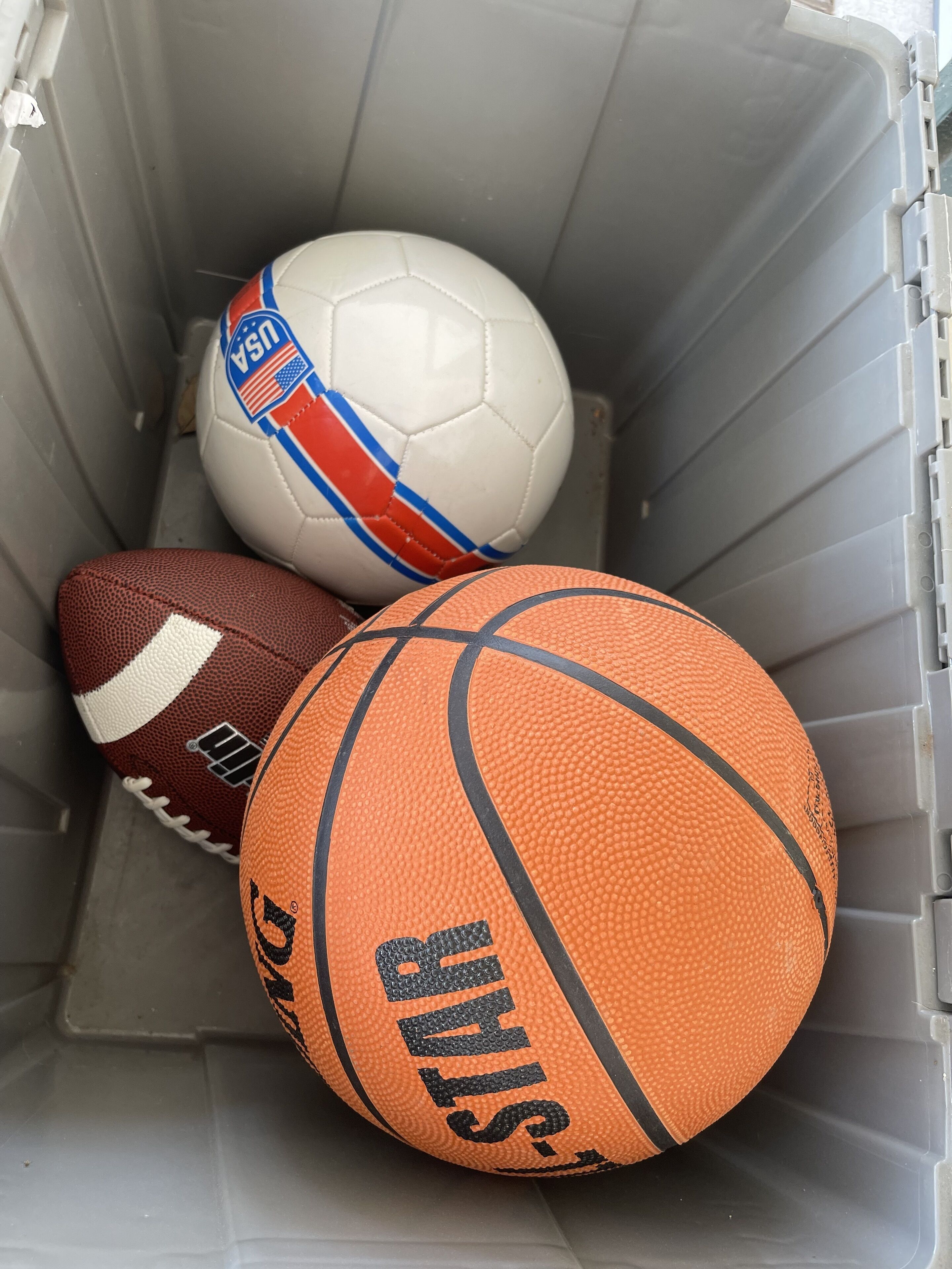 Un récipient de rangement contenant trois ballons de sport : un ballon de football américain, un ballon de basket-ball et un ballon de football.