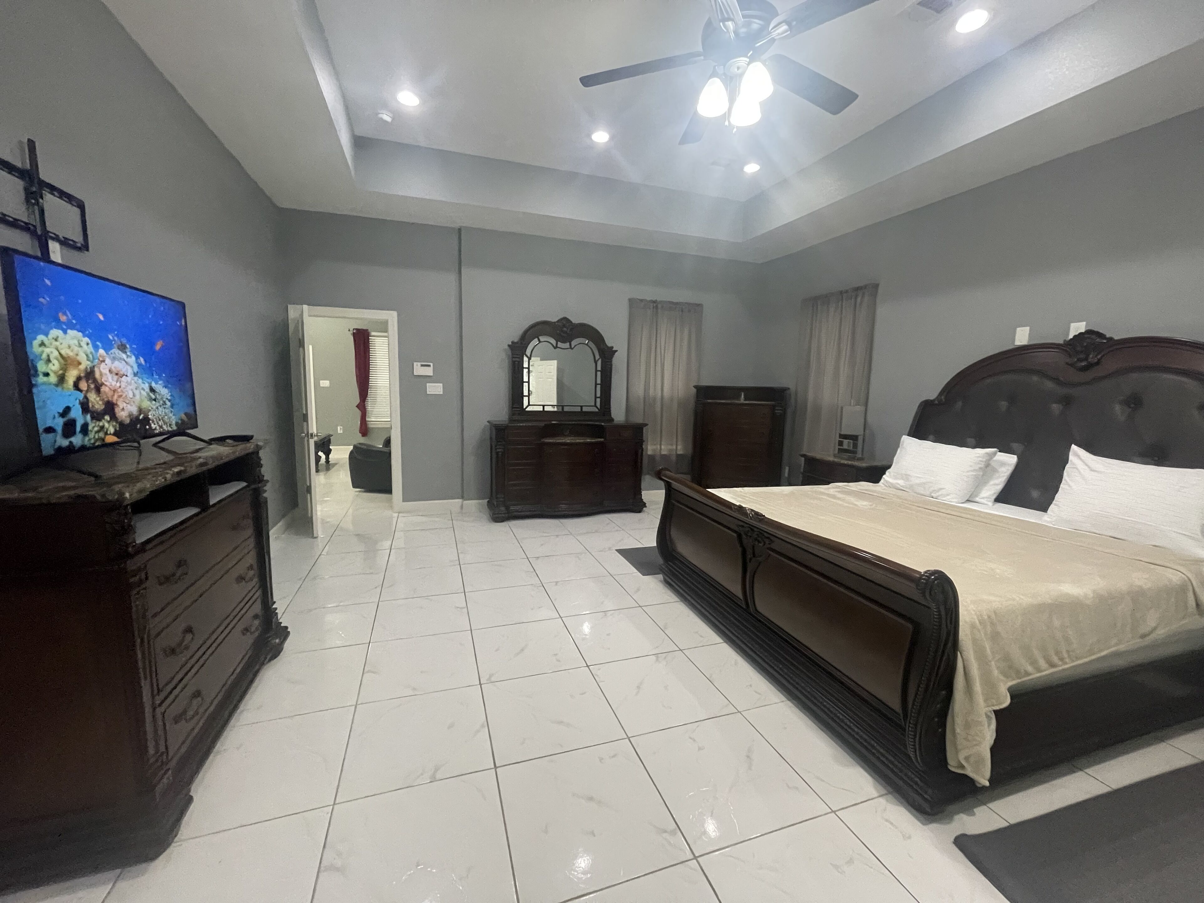 Ein Schlafzimmer mit Bett, Schminktisch, Fernseher, Deckenventilator und einem durch eine offene Tür einsehbaren Badezimmer.