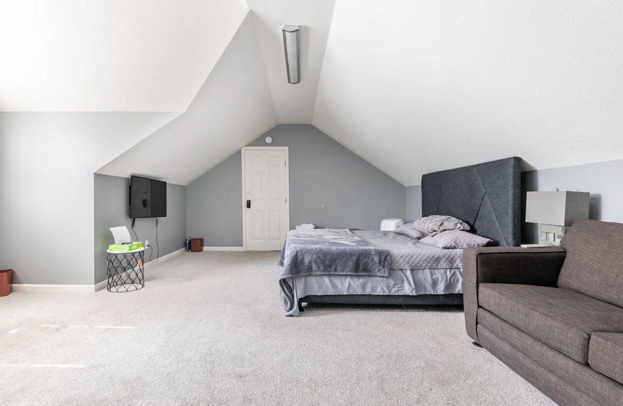 A bedroom with a bed, a grey sofa, a TV mounted on the wall, and a door.