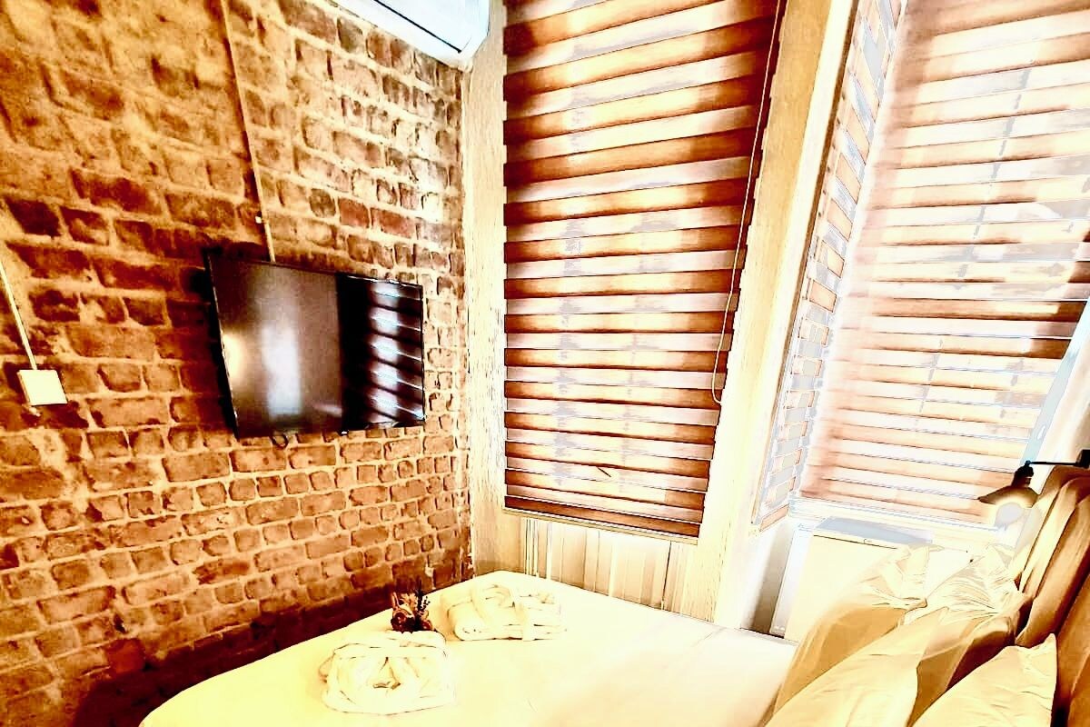Ein Zimmer mit Backsteinwand, einem Bett mit weißer Bettwäsche, einem Holztisch mit Teller und Flasche sowie einem an der Wand montierten Fernseher.