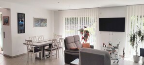 Una sala de estar moderna con comedor, sofá, televisor y un ventanal con persianas.