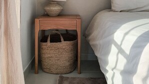 Une chambre à coucher avec un lit, une table de chevet avec une lampe, un panier en osier et un rideau.