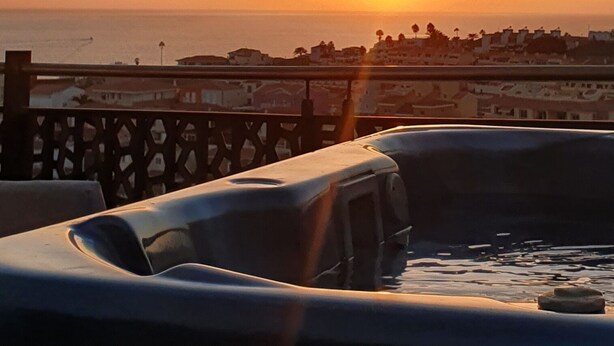 Une vue sur le coucher de soleil depuis un balcon doté d’une balustrade, donnant sur une zone côtière avec des bâtiments et la mer.