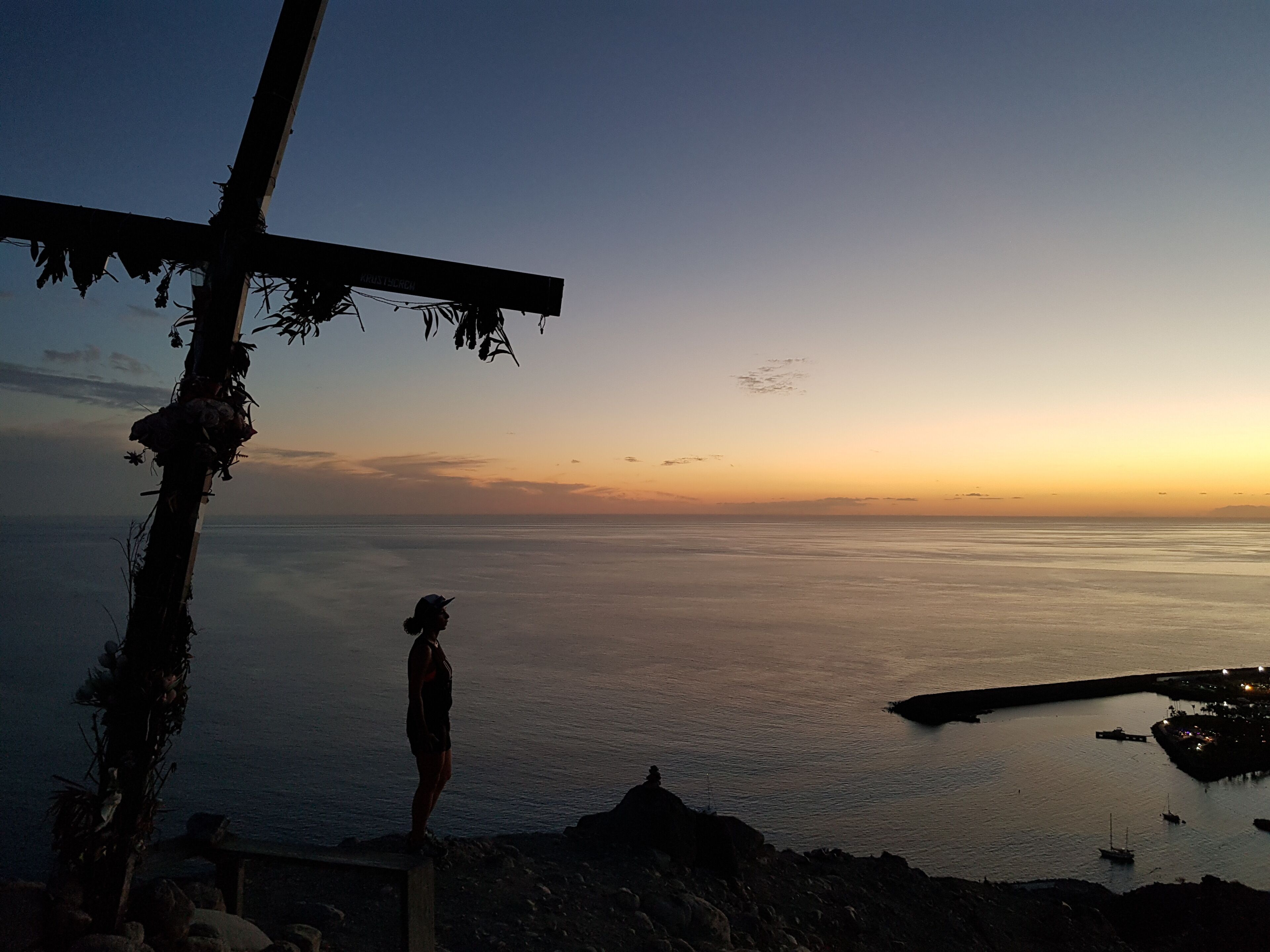 Une vue sur le coucher de soleil sur la côte, avec une croix et une personne debout sur un promontoire rocheux.