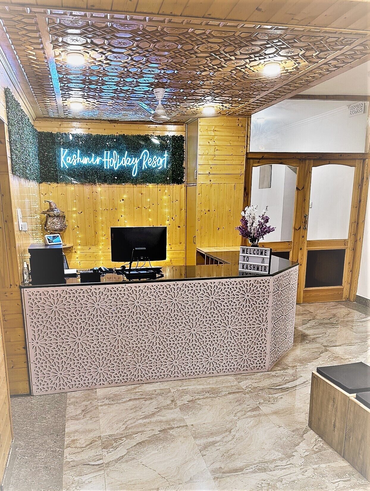 A reception area with a wooden reception desk, a neon sign reading "Kashmir Holiday Resort," and a "WELCOME TO TASHMIR" sign on the counter.